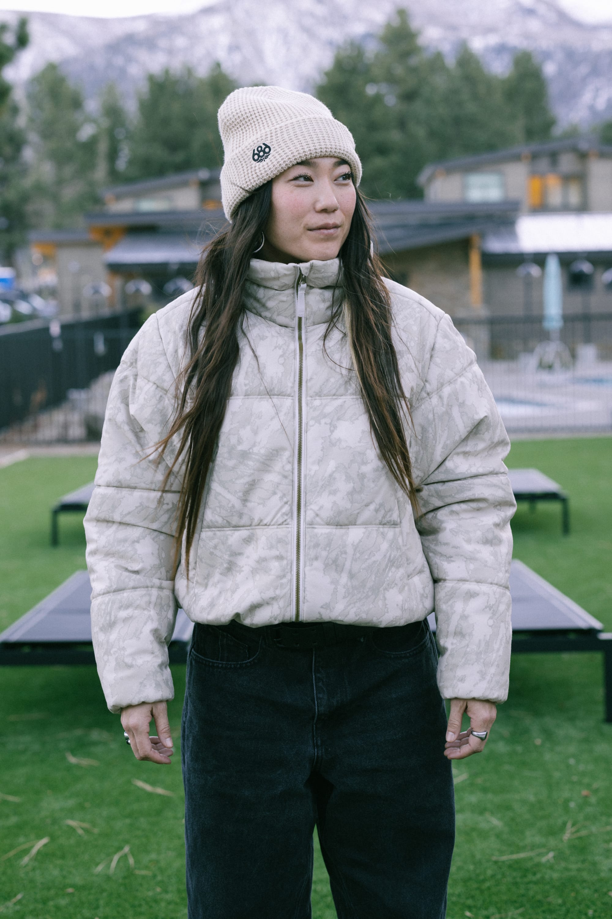 A woman wearing the 686 Women’s Gracious Puffer Jacket and a beige beanie stands outdoors on green grass, with mountains and buildings in the background.