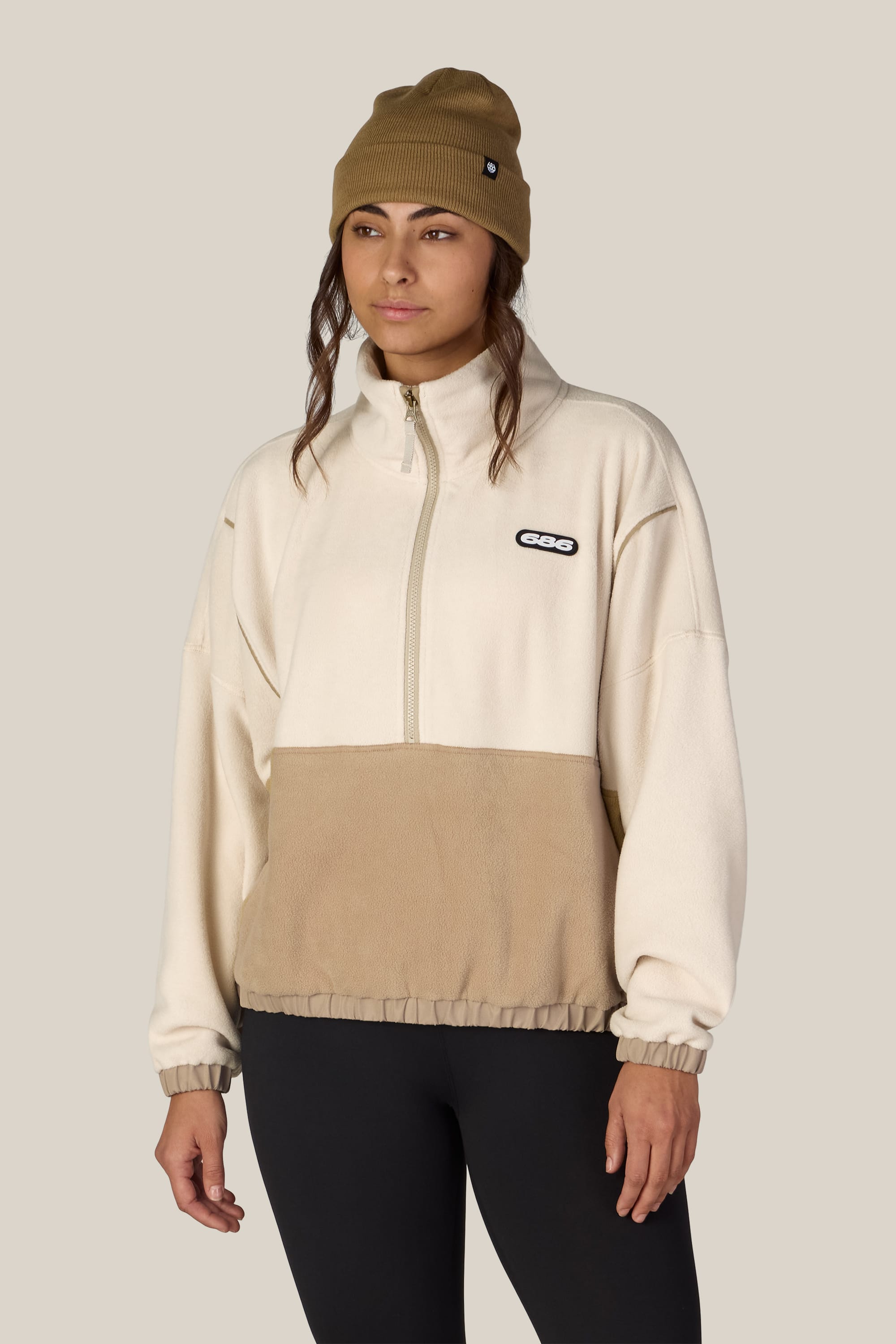 A woman wearing the 686 Women’s Chimay Fleece Anorak, a cozy cream and tan zip-up pullover made from 100% polyester by 686, pairs it with a beige beanie and black leggings as she stands against a plain light background, highlighting the casual versatility of this women’s jacket.