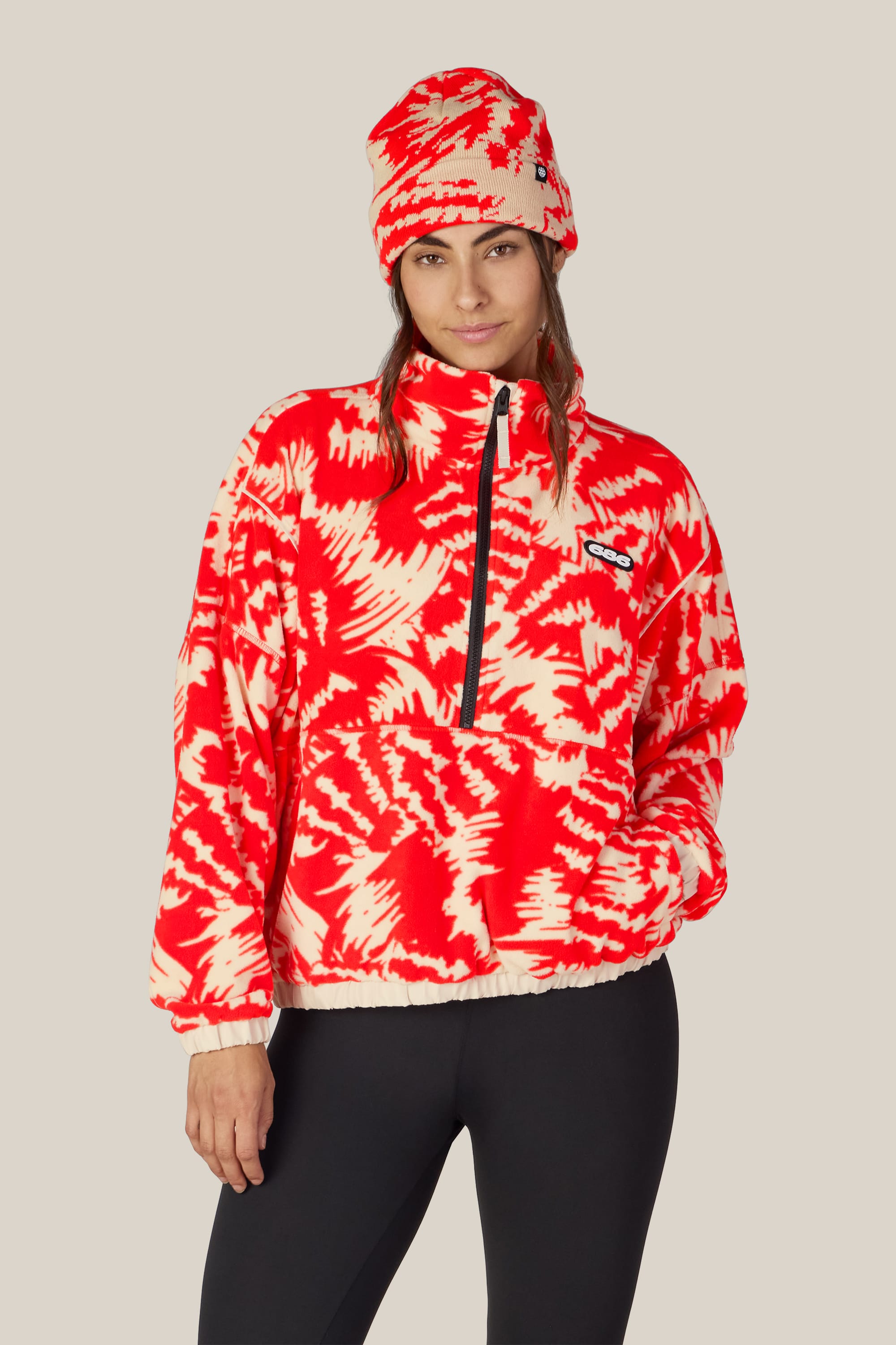 A person wearing the 686 Women’s Chimay Fleece Anorak—a red and cream patterned pullover from 686—paired with a matching beanie and black leggings, stands facing forward against a plain light beige background. Crafted from 100% polyester for casual comfort, this cozy anorak adds a modern touch reminiscent of a women’s jacket.