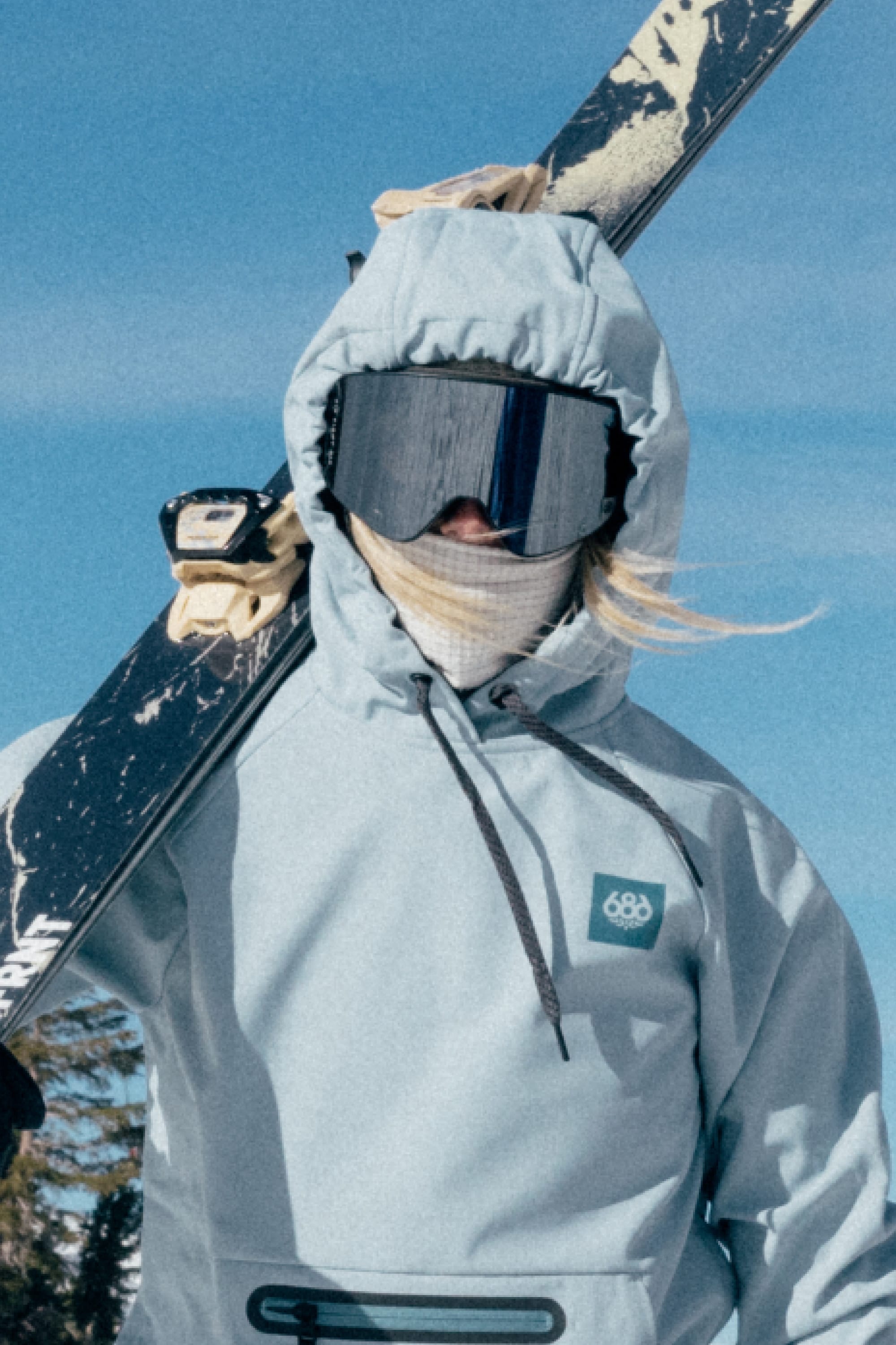 A person in light blue winter gear and goggles, wearing the 686 Mission Grid™ Balaclava by 686, holds skis over their shoulder while standing outside on a snowy day.