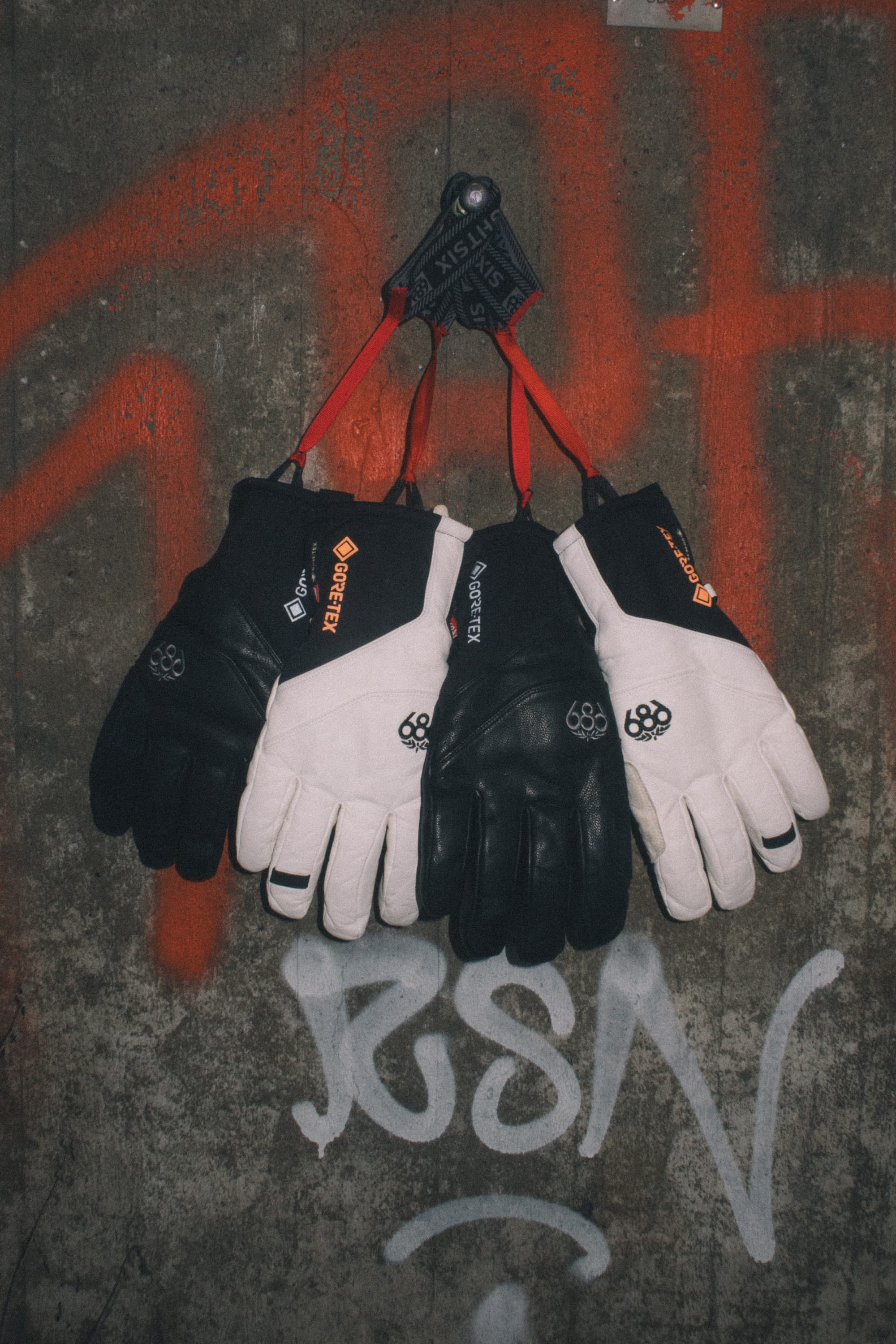 Four pairs of black and white winter gloves, including the 686 Men’s GORE-TEX® Stronghold Leather Glove by 686 featuring durable leather palms, hang on a hook against a concrete wall with red and white graffiti.