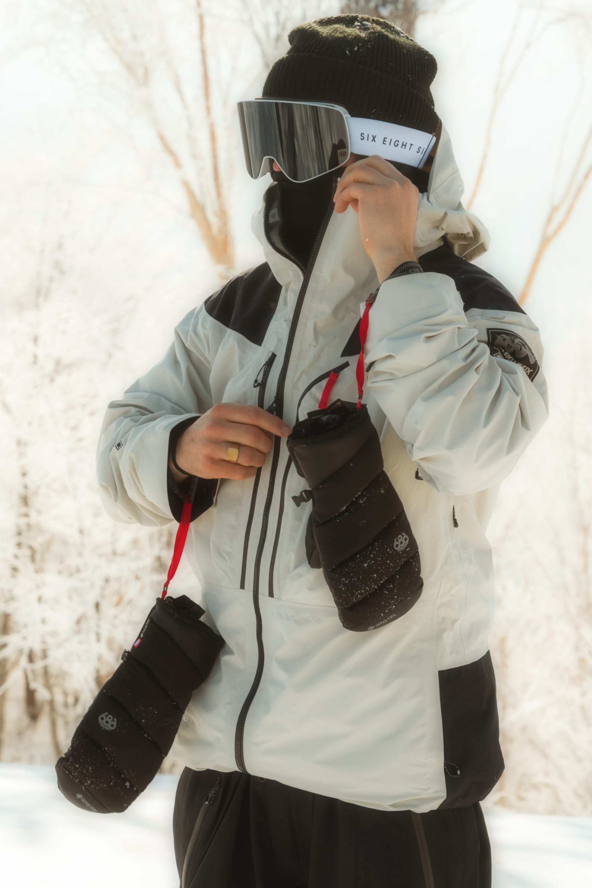 A person in winter gear and goggles stands outdoors in the snow, holding a pair of black insulated bottle holders with red straps, while wearing the 686 Men’s GORE-TEX® Buffer Insulated Mitt to keep their hands warm.
