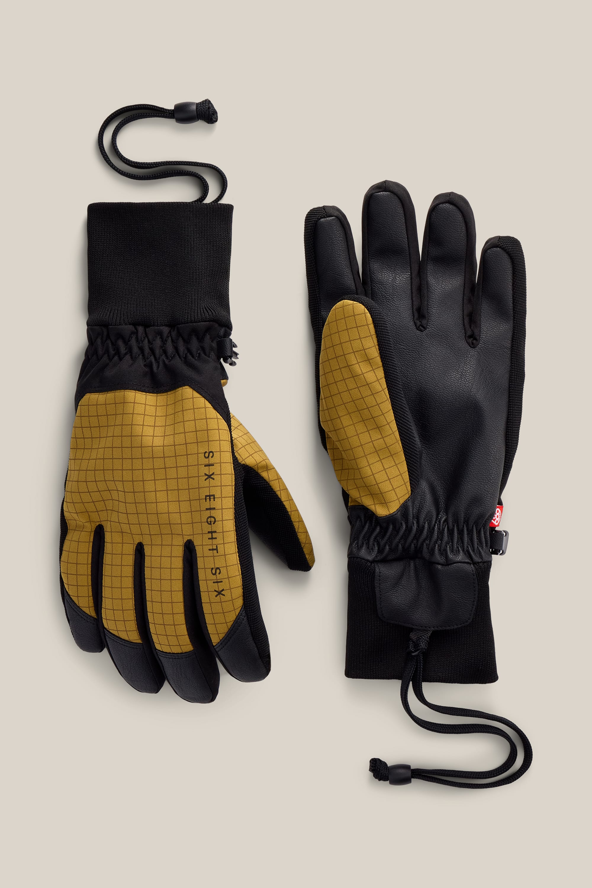 A pair of 686 Men’s Crush Insulated Gloves in yellow and black with a grid pattern, featuring black PU palms, adjustable wrist cuffs, and attached elastic cords, displayed on a neutral background.
