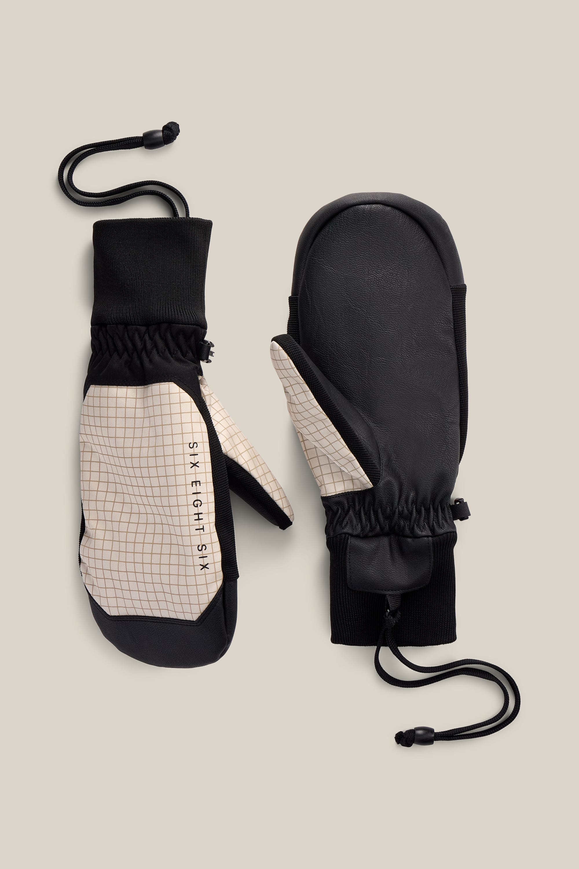 A pair of 686 Men’s Crush Insulated Mitts featuring black palms and beige checkered fabric on top, elastic wrists, and adjustable wrist cords. The brand logo “686” is printed on the side of one mitten, adding a stylish touch to this versatile insulated poly mitt design.