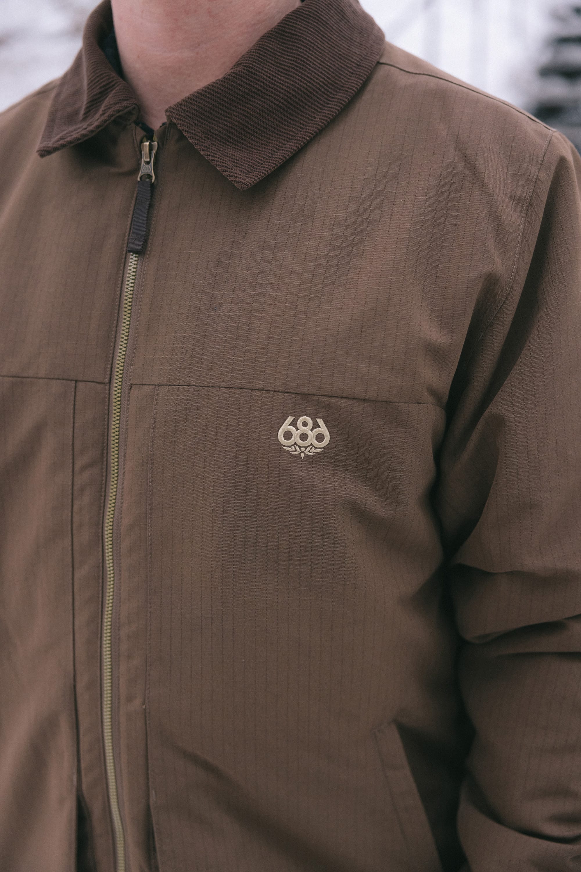 A person wearing the 686 Men’s Camber Insulated Jacket in brown, featuring a corduroy collar and a small 686 logo on the chest, stands outside. Only the upper torso is visible.