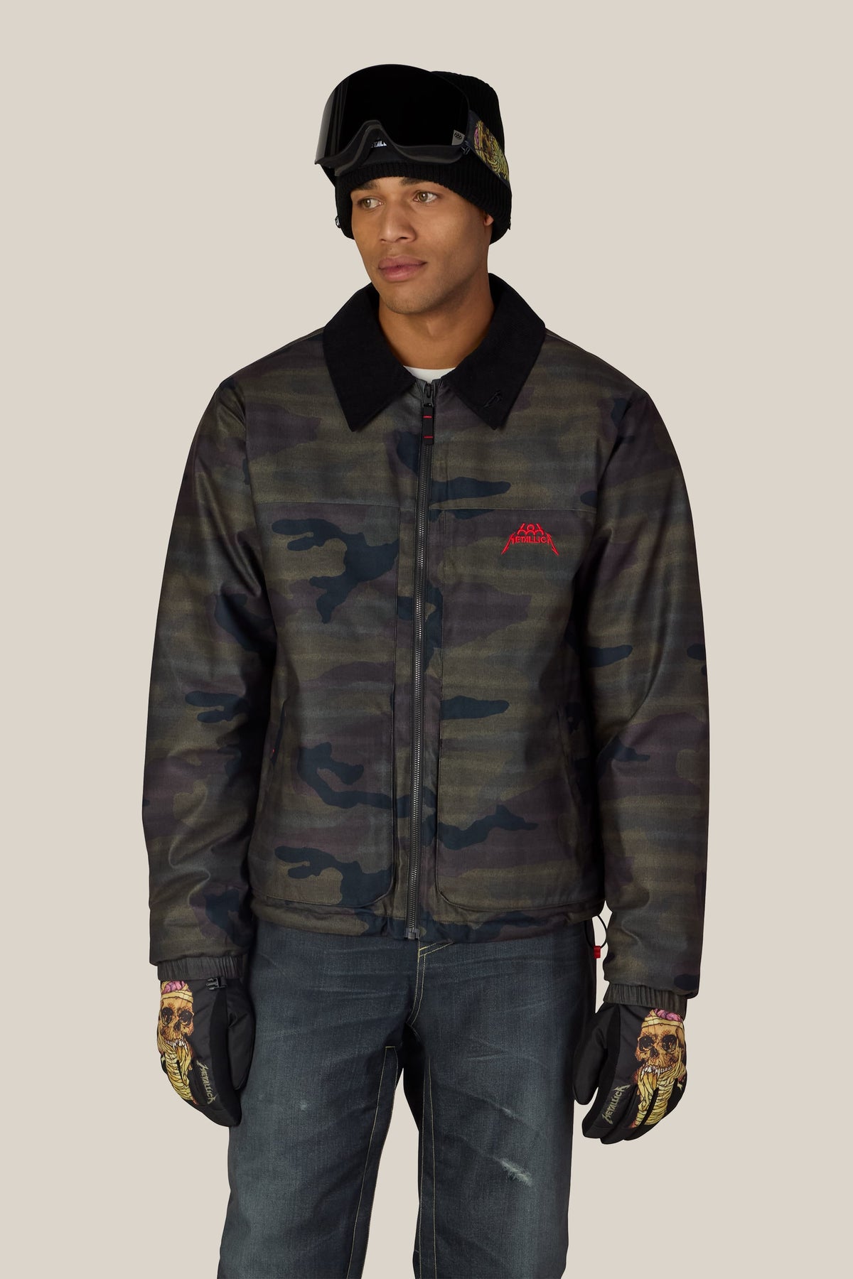 A person wearing a 686 Men’s Metallica Caim Jacket with a black collar, black beanie, ski goggles, and skull-design gloves.