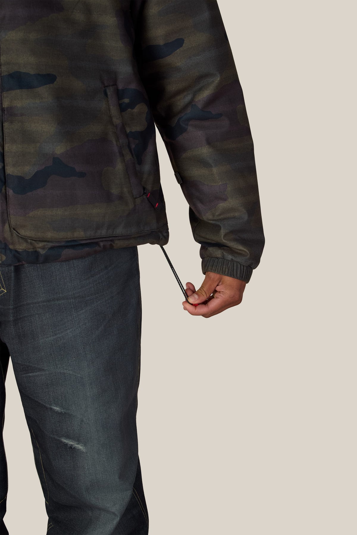 A person wearing a camo-print 686 Men’s Metallica Caim Jacket and dark jeans pulls on the adjustable drawstring at the hem.