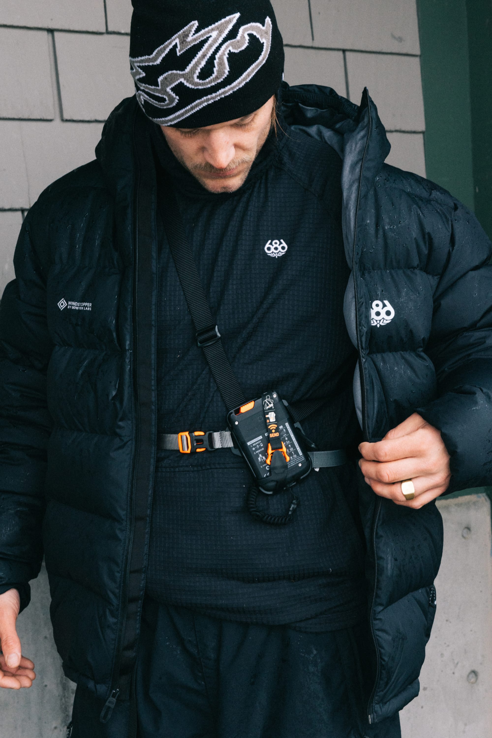 Dressed in a black puffer jacket, black pants, and a black beanie featuring a white flame design, the person looks down as they adjust their jacket. For added warmth, they’ve layered the 686 Men’s Mission Grid™ Raglan Hoody by 686 underneath. An orange-accented crossbody bag completes the look.