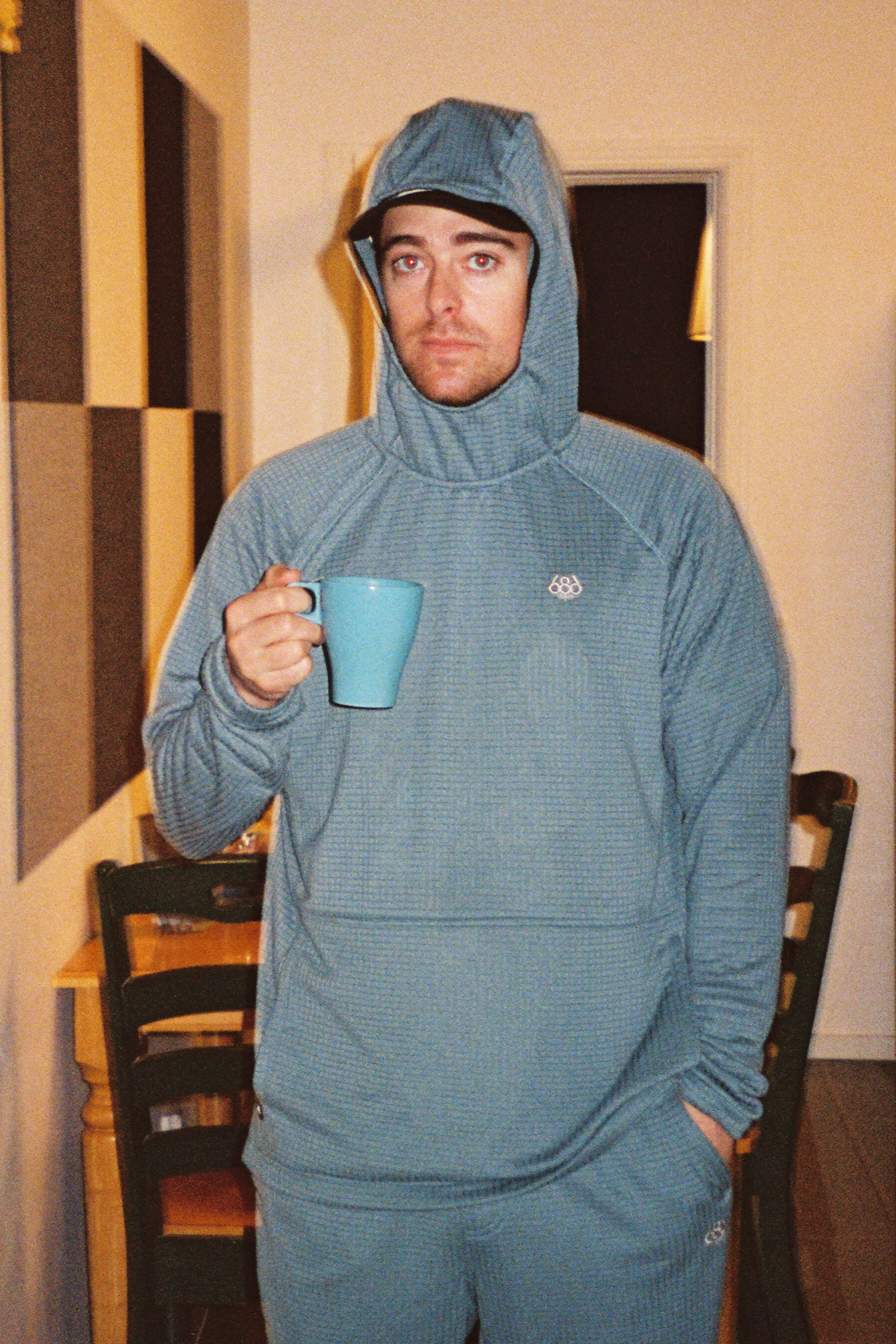 A man wearing a blue 686 Men’s Mission Grid™ Raglan Hoody stands indoors, holding a blue mug and looking at the camera with a neutral expression. Chairs and a table are visible in the background.