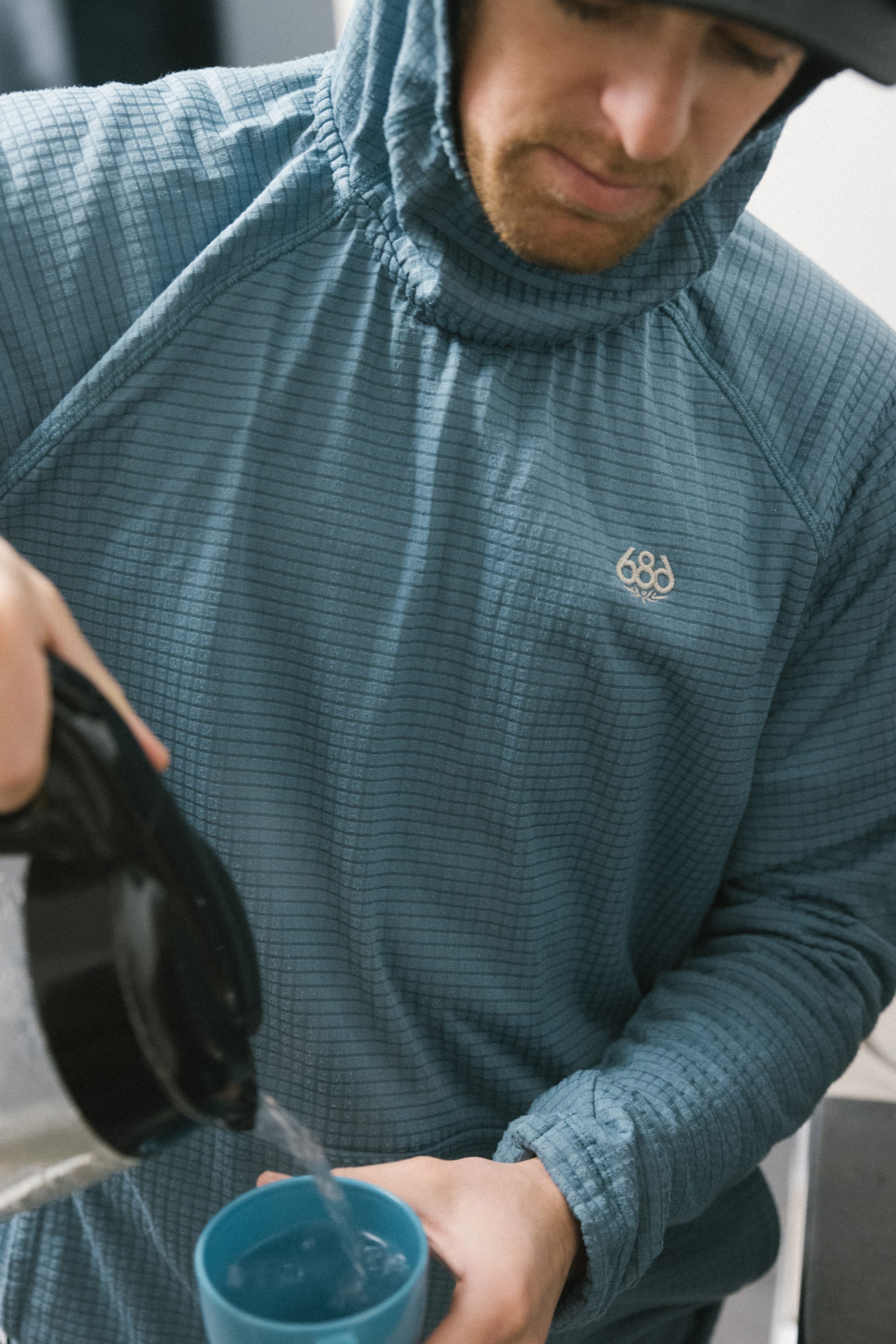 Wearing the 686 Men’s Mission Grid™ Raglan Hoody by 686, a person pours hot water from a black kettle into a blue mug, with their face partially visible and head slightly tilted down.