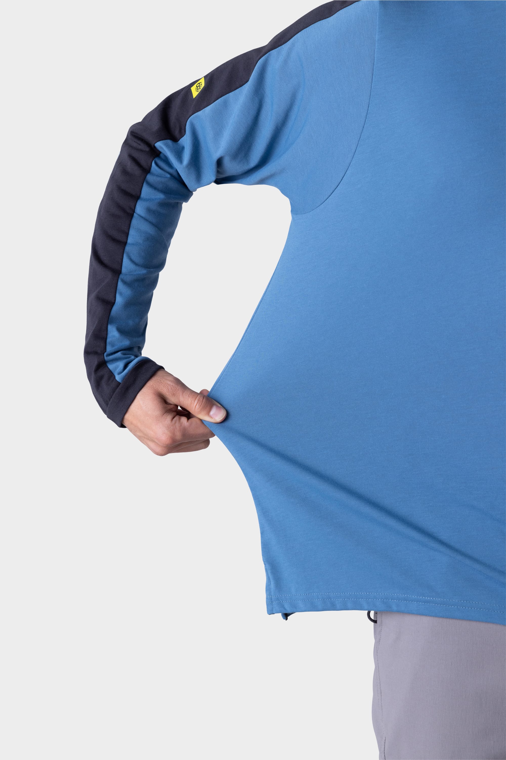 A person stretches the fabric of a blue 686 Mens Rival Long Sleeve Bike Jersey with one hand, demonstrating its flexibility against a plain white background.