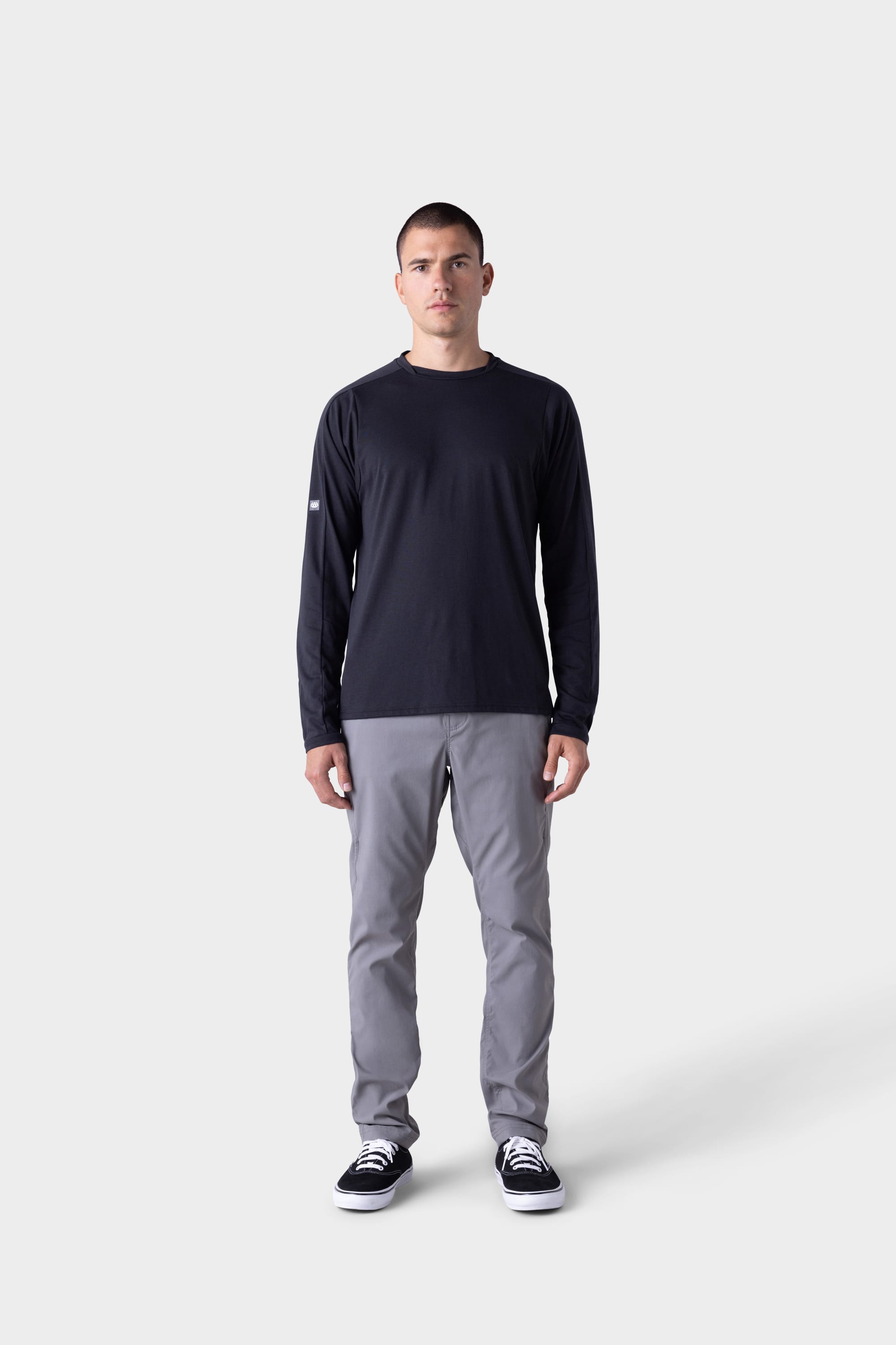 A man faces forward against a plain light gray background, wearing the 686 Mens Rival Long Sleeve Bike Jersey, light gray pants, and black-and-white sneakers.