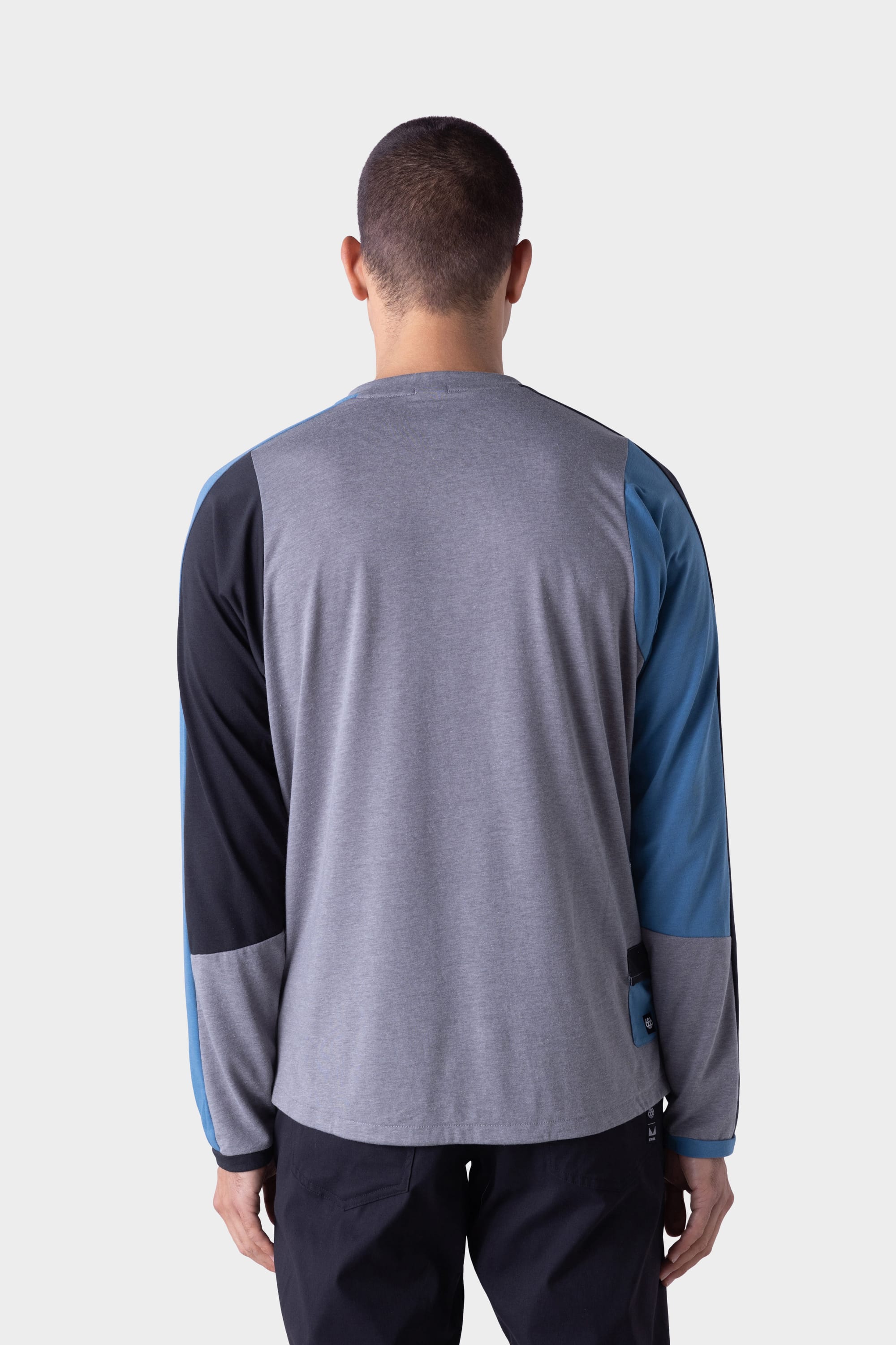 A person stands with their back to the camera, wearing the 686 Mens Rival Long Sleeve Bike Jersey by 686 in gray, black, and blue color-blocked sleeves, paired with black pants against a plain light background.