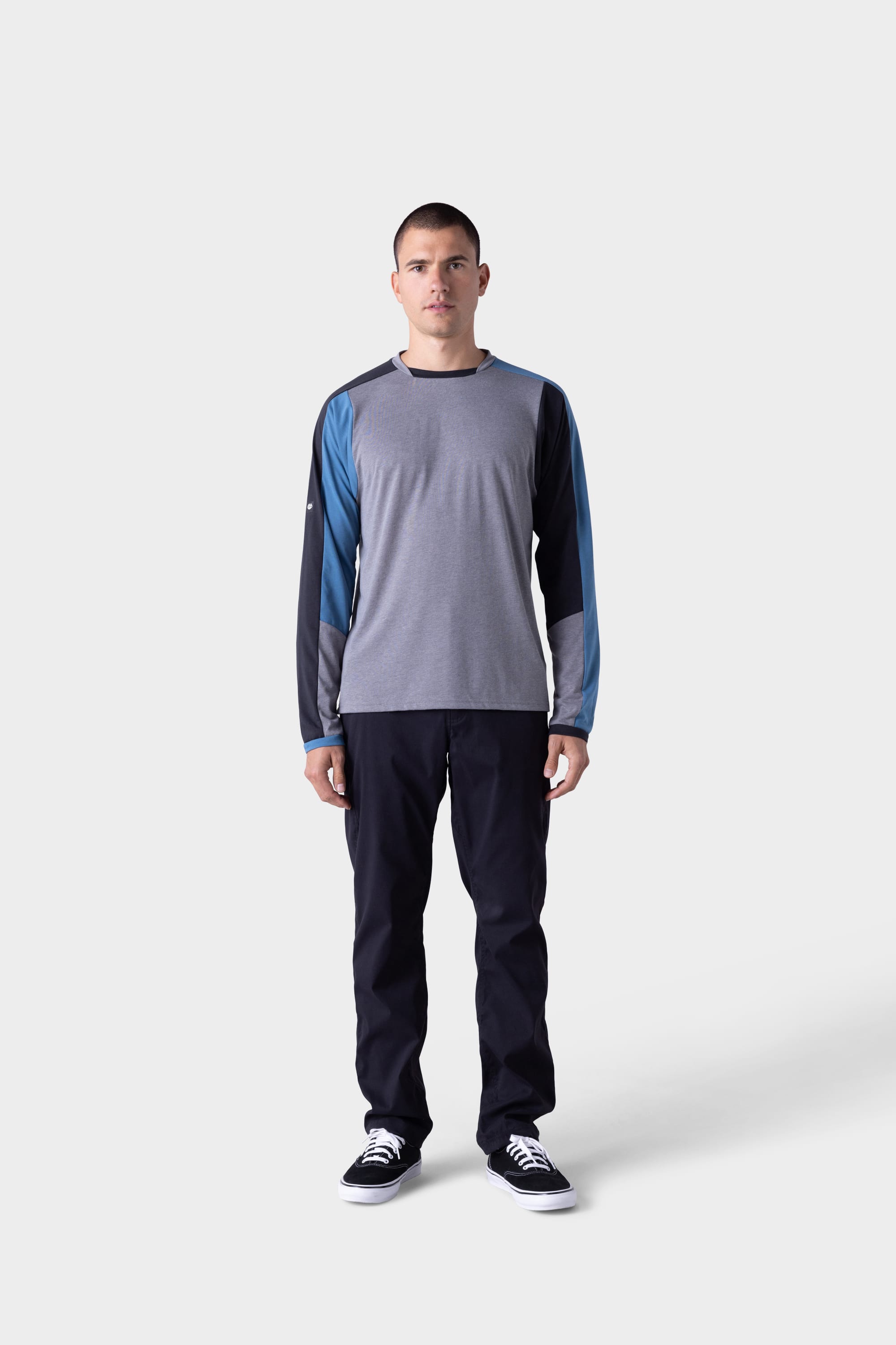 A man faces forward against a plain light gray background, wearing the 686 Mens Rival Long Sleeve Bike Jersey by 686 in shades of gray and blue, paired with dark pants and black-and-white sneakers.