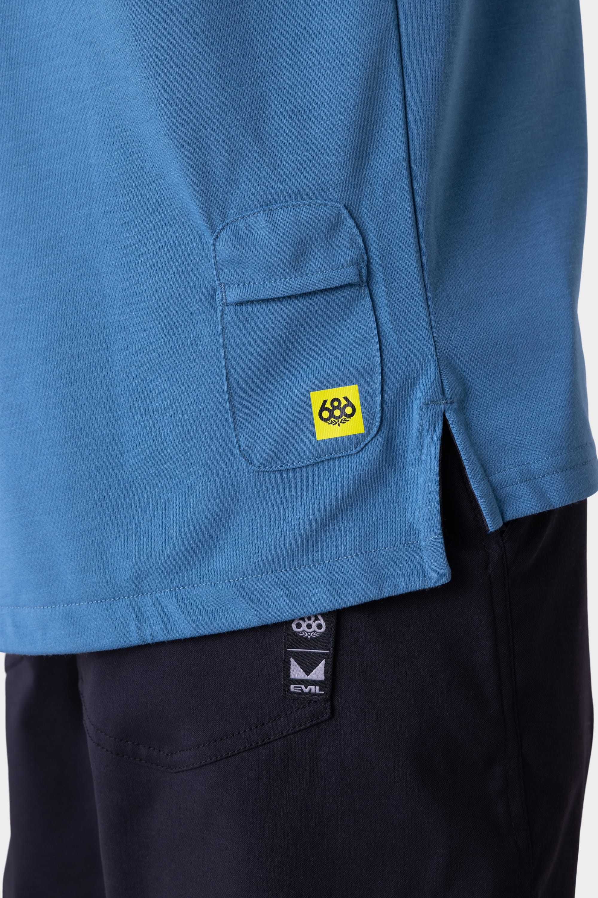 Close-up of the 686 Mens Rival Long Sleeve Bike Jersey in blue, showing a small pocket and yellow 686 logo above black pants with a visible black and white logo tag.