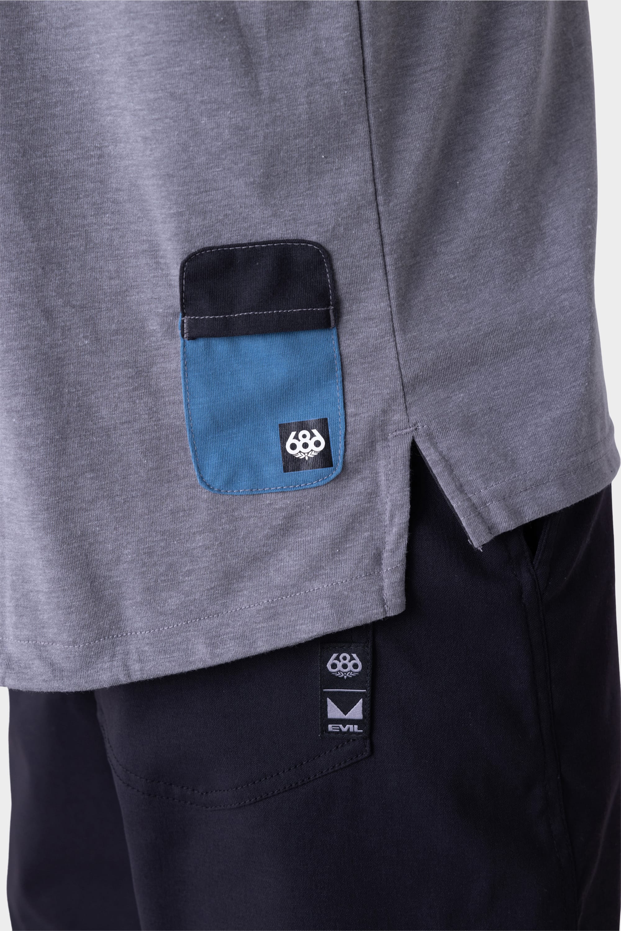 Close-up of the 686 Mens Rival Long Sleeve Bike Jersey in gray, featuring a small blue and black pocket detail with a logo near the hem, paired with black pants displaying a logo tag.