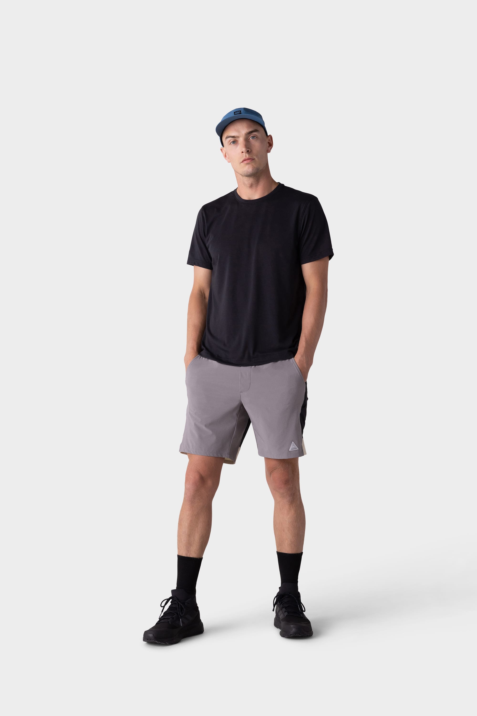 A man stands with hands in pockets, wearing a blue cap, black t-shirt, 686 Mens ATP Stretch Performance Short by 686 in gray, black socks, and black shoes against a plain white background.